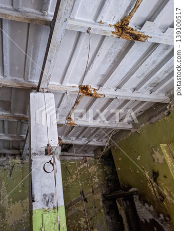 The close view shows a large supporting column and a damaged ceiling of an ancient industrial building. Rust and wear reveal the history of the space and its long neglect. The close view shows a large supporting column and a damaged ceiling of an ancient industrial building. Rust and wear reveal the history of the space and its long neglect. 139100517