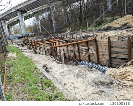 Workers are reinforcing a deep trench at a construction site. Wooden planks and metal pipes are in place along the trench. A highway overpass is visible above the site. Workers are reinforcing a deep trench at a construction site. Wooden planks and metal pipes are in place along the trench. A highway overpass is visible above the site. 139100518