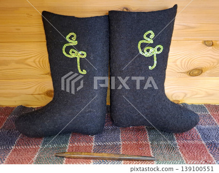 A pair of traditional Russian black felt boots sits on a woven rug. The boots are decorated with green sequins. The background shows warm wooden walls. This is a cozy indoor location. A pair of traditional Russian black felt boots sits on a woven rug. The boots are decorated with green sequins. The background shows warm wooden walls. This is a cozy indoor location. 139100551