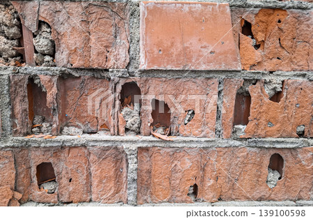 A close up of a red brick wall that is old and damaged. Erosion has caused significant wear, creating holes and crumbling areas in the bricks. 139100598