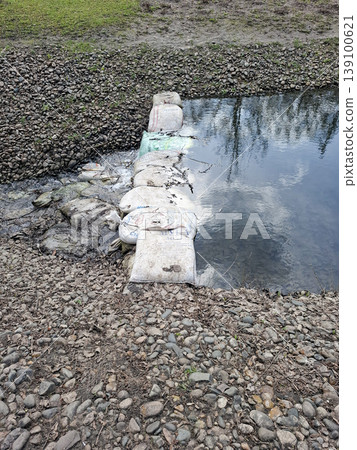 A small dam is built across a narrow stream using white sandbags. The setting is a rural or park area with visible stones and grass nearby. Water flows over the dam. 139100621