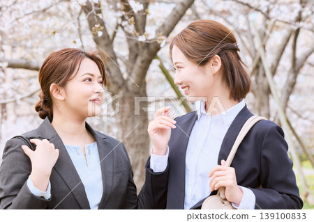 Two young businesswomen in suits, new employees, chatting under a row of cherry trees—a spring scene where they smile and talk. 139100833