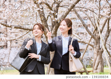 Two female new employees in suits enjoying cherry blossom viewing under a row of cherry trees - a spring commute scene. Two female new employees in suits enjoying cherry blossom viewing under a row of cherry trees - a spring commute scene. 139100834