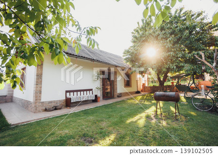 Sunlit Backyard With Bicycle And Barbecue, Warm Golden Light Filtering Through Trees Over Grass And Bench Mood Evokes Family Leisure, Evening Picnic, Relaxed Summer Lifestyle Scenes. Sunlit Backyard With Bicycle And Barbecue, Warm Golden Light Filtering Through Trees Over Grass And Bench Mood Evokes Family Leisure, Evening Picnic, Relaxed Summer Lifestyle Scenes. 139102505