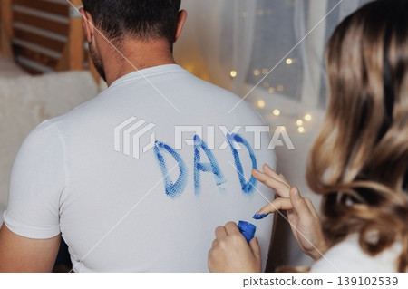 Man In White Shirt Painted DAD, Woman Applying Blue Paint Heart On Lower Back, Cozy Room With Fairy Lights, Wooden Floor, Casual Jeans, Playful Intimate Moment, DIY Gift Preparation Man In White Shirt Painted DAD, Woman Applying Blue Paint Heart On Lower Back, Cozy Room With Fairy Lights, Wooden Floor, Casual Jeans, Playful Intimate Moment, DIY Gift Preparation 139102539