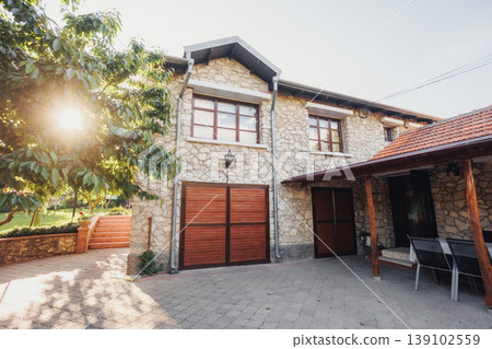 Textured Stone Garage And Sunlit Patio With Seating, Wooden Doors And Shadowed Porch Area, Rustic Materials, Detailed Surfaces And Intimate Exterior Corner Perfect For Architecture Textured Stone Garage And Sunlit Patio With Seating, Wooden Doors And Shadowed Porch Area, Rustic Materials, Detailed Surfaces And Intimate Exterior Corner Perfect For Architecture 139102559