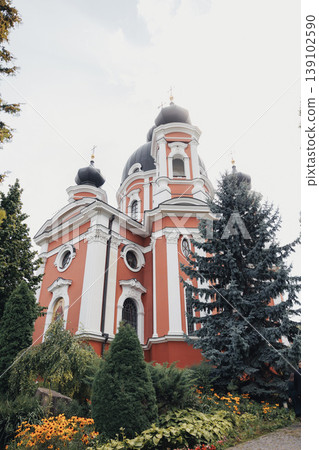 Ornate Red Orthodox Church Exterior Framed By Evergreens, Baroque Towers With Cupola Silhouettes Rising Above Manicured Gardens, Textured Stucco Facade, Decorative Cornices, Tranquil Ornate Red Orthodox Church Exterior Framed By Evergreens, Baroque Towers With Cupola Silhouettes Rising Above Manicured Gardens, Textured Stucco Facade, Decorative Cornices, Tranquil 139102590
