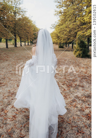 Silken Wedding Dress Amidst Autumn Foliage. Bride In Delicate Dress Walks Through Peaceful Leafy Environment. Graceful Bride In Airy Dress Moves Between Trees Under Soft Fall Sunlight 139102615