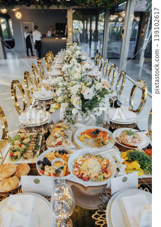 Lavish Banquet Table With Extensive Plated Courses And Towering Floral Runners, Gold Chairs, Ornate Tableware, Abundant Salads And Entrees Staged For Gala Reception In Upscale Venue 139102617