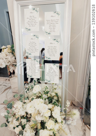 Mirror Seating Chart With White Florals, Handlettered Calligraphy On Glass Panels, Gold Frame Anchored Among Lush Roses And Peonies, Marble Floor Reflection, Scattered Petals, Soft Natural 139102618