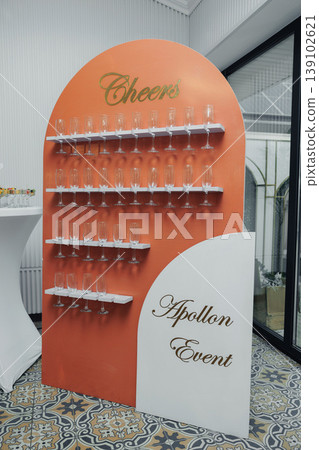 Champagne Glass Display Wall Featuring Rows Of Clear Flutes On Orange Curved Arch, Cheers Header With Apollon Event Script, Elegant Reception Setup For Corporate Launch, Patterned Tile 139102621