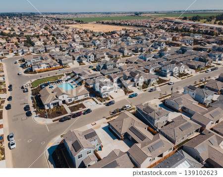 Aerial Family Suburb With Community Pool At Center Showing Homes Circling Pool, Landscaped Common Area, Swim Lanes, Sidewalks, Parked Cars And Bright Summer Light Over Fields. 139102623