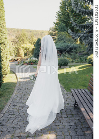 Bride In Veil Walking Garden Path, Caucasian Bride Moving Away Along Stone Walkway Through Manicured Greenery, Long Train Flowing, Soft Backlight, Contemplative Mood And Elegant Gown Bride In Veil Walking Garden Path, Caucasian Bride Moving Away Along Stone Walkway Through Manicured Greenery, Long Train Flowing, Soft Backlight, Contemplative Mood And Elegant Gown 139102633