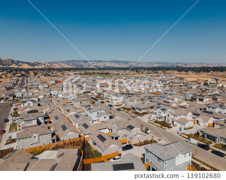 Aerial New Housing Development With Mountain Backdrop Showing Dense Rooftops, Construction Patches, Fenced Yards, Grid Streets, Ongoing Site Preparation And Distant Peaks Under Clear Sky. 139102680