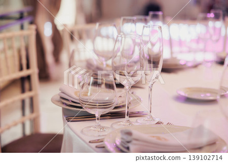 Elegant Table Setting With Glassware, Soft Pink Lighting, Neatly Folded Napkins On White Plates, Crystal Wine Glasses Aligned, Blurred Banquet Chairs In Background, Intimate Romantic Elegant Table Setting With Glassware, Soft Pink Lighting, Neatly Folded Napkins On White Plates, Crystal Wine Glasses Aligned, Blurred Banquet Chairs In Background, Intimate Romantic 139102714