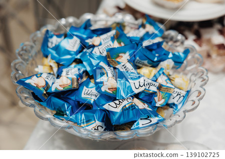 Glass Bowl Filled With Wrapped Candies In Blue Packaging, Glossy Wrappers Catching Light, Closeup Shot On Party Table With Blurred Desserts And Plates In Background, Festive Catering Display 139102725