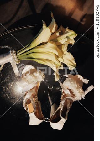 Overhead Bridal Shoes With Calla Bouquet Reflected On Dark Glass Surface, Long Stems And Crisp Petals Framing Delicate Heels, Cinematic Warm Tones And Moody Shadows Create Romantic Tableau. 139102745