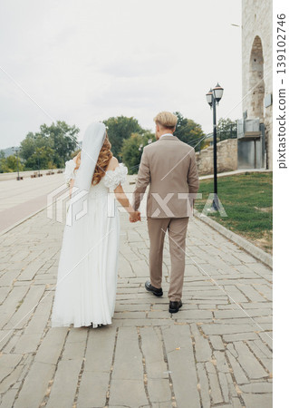Bride And Groom Walking Along Cobblestone Street, Caucasian Couple Moving Through Historic Urban Setting, Tailored Suit And Flowing Dress, Casual Conversation And Refined Atmosphere. Bride And Groom Walking Along Cobblestone Street, Caucasian Couple Moving Through Historic Urban Setting, Tailored Suit And Flowing Dress, Casual Conversation And Refined Atmosphere. 139102746