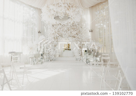 Bright White Floral Ceremony Hall With Towering Flower Arch And Minimalist Altar, Sunlight Floods Through Sheer Drapery Onto Glossy Aisle, Rows Of Simple Chairs Frame Cascading Blossoms, 139102770