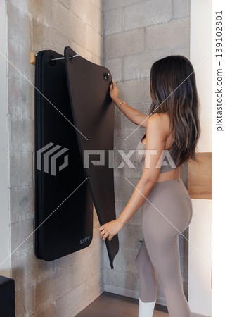 Woman Folding Wallmounted Treadmill Panel, Preparing Home Gym Setup With Careful Adjustment Of Straps, Concrete Wall Backdrop, Beige Activewear, Back View, Compact Storage Solution, Calm 139102801
