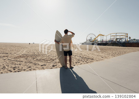 Surfer On Sunlit Boardwalk Holding Surfboard, Caucasian Male Gazing Toward Distant Pier With Long Shadow Across Pavement, Warm Hazy Light, Empty Sand And Calm Ocean, Relaxed Contemplative Surfer On Sunlit Boardwalk Holding Surfboard, Caucasian Male Gazing Toward Distant Pier With Long Shadow Across Pavement, Warm Hazy Light, Empty Sand And Calm Ocean, Relaxed Contemplative 139102804