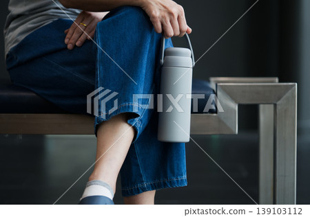 Sleek Matte Grey Insulated Reusable Water Bottle with Handle, held while sitting on a Modern Bench Sleek Matte Grey Insulated Reusable Water Bottle with Handle, held while sitting on a Modern Bench 139103112