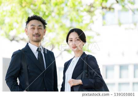 Amidst the fresh greenery, smiling businessmen and women stand in front of an outdoor building, projecting an image of trust and career success, looking directly at the camera with smiles. Amidst the fresh greenery, smiling businessmen and women stand in front of an outdoor building, projecting an image of trust and career success, looking directly at the camera with smiles. 139103801