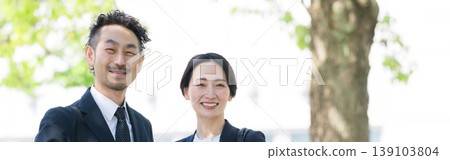 Amidst the fresh greenery, a smiling business couple in front of an outdoor building, conveying an image of trust and career success, with smiling faces and a banner. 139103804