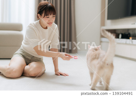Happy asian woman sitting on living room floor at home taking joyful break from work and study using technology to feed fluffy cat smiling warmly playing with cute domestic pet Happy asian woman sitting on living room floor at home taking joyful break from work and study using technology to feed fluffy cat smiling warmly playing with cute domestic pet 139104491