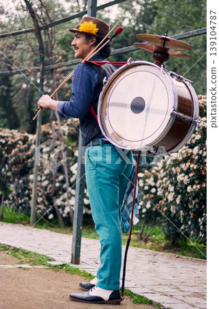 Adult man with a mustache, dressed formally and colorfully, posing and playing a bass drum on his back, Chilean chinchinero, street musician who plays the bass drum and dances in the streets Adult man with a mustache, dressed formally and colorfully, posing and playing a bass drum on his back, Chilean chinchinero, street musician who plays the bass drum and dances in the streets 139104787