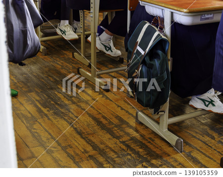 A green school bag hangs beside a desk in a classroom in a wooden school building. A green school bag hangs beside a desk in a classroom in a wooden school building. 139105359