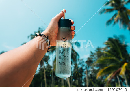 Hand holding a reusable water bottle against a background of tropical palm trees and clear blue sky. 139107043