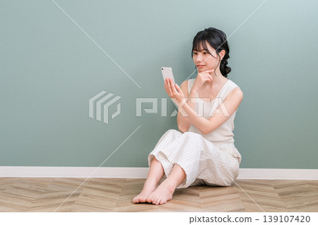 A young Asian woman thinking and considering while looking at her smartphone at home (worried, serious, considering, planning) A young Asian woman thinking and considering while looking at her smartphone at home (worried, serious, considering, planning) 139107420