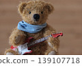 A teddy bear holding a guitar; yellow bear, red guitar; close-up. 139107718