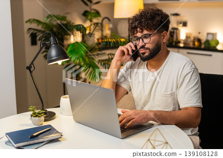 Arabian man freelancer works on laptop and talks on smartphone during remote job evening indoors Arabian man freelancer works on laptop and talks on smartphone during remote job evening indoors 139108128