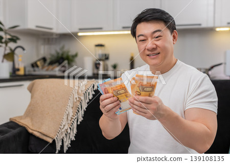 Asian man counts cash euros smiles makes money sign and plans invest purchase bonus salary savings Asian man counts cash euros smiles makes money sign and plans invest purchase bonus salary savings 139108135
