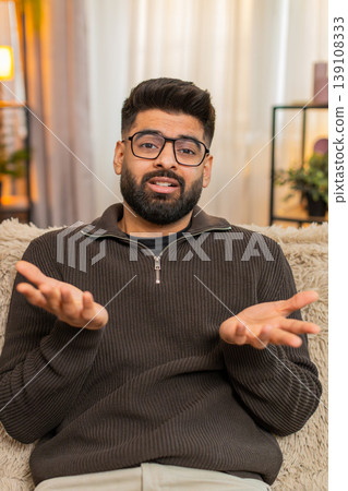 Indian man at sofa spreads hands in confusion and surprise at home, asking what with puzzled face 139108333