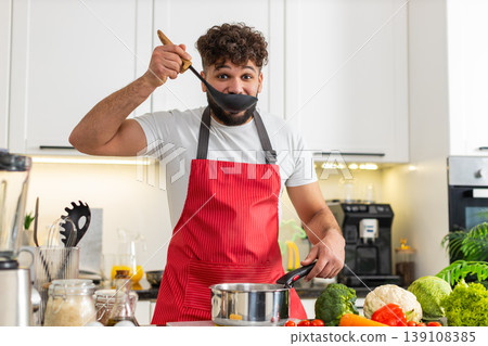 Arabian young adult man smiling stirring soup pot on stove in home kitchen for healthy dinner recipe Arabian young adult man smiling stirring soup pot on stove in home kitchen for healthy dinner recipe 139108385