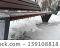 A park bench covered in ice and hanging icicles sits in snow on a winter day with other benches and trees in the background cold frosty weather urban scenery travel concept 139108818