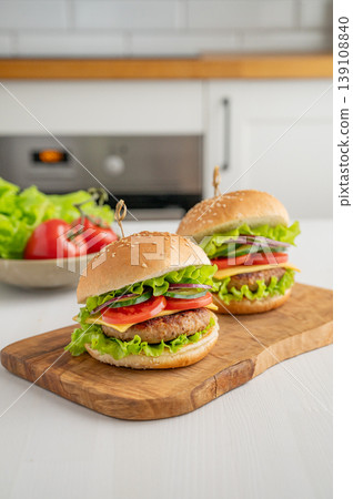 Two beef burgers with tomato, cheese, and lettuce on a cutting board on a light table Two beef burgers with tomato, cheese, and lettuce on a cutting board on a light table 139108840