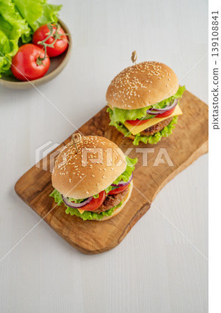 Two beef burgers with tomato, cheese, and lettuce on a cutting board on a light wooden background Two beef burgers with tomato, cheese, and lettuce on a cutting board on a light wooden background 139108841