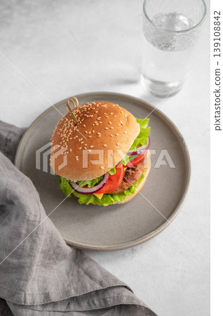 Beef burger with tomato, cheese, and lettuce on a plate on a kitchen table with glass of water  139108842