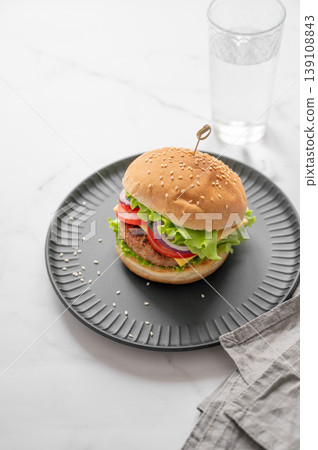 Beef burger with tomato, cheese, and lettuce on a plate on a marble background with glass of water 139108843