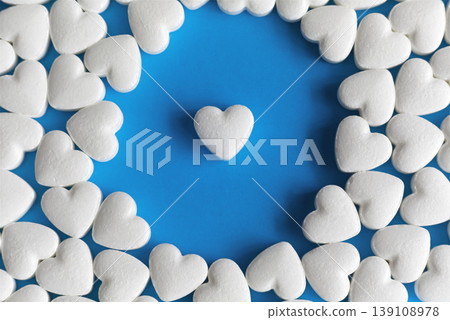 White heart-shaped pill in the center on a blue background surrounded by a halo of other heart pills, macro shot of heart-shaped pills on a blue background for content about heart health. White heart-shaped pill in the center on a blue background surrounded by a halo of other heart pills, macro shot of heart-shaped pills on a blue background for content about heart health. 139108978
