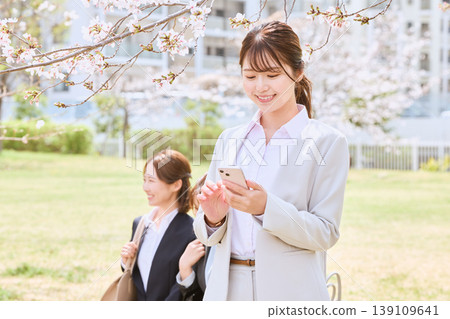 A young female business person with a smile operating her smartphone under cherry blossoms – an image of a new spring life. A young female business person with a smile operating her smartphone under cherry blossoms – an image of a new spring life. 139109641