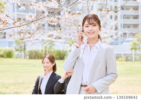 A young businesswoman talking on her smartphone under cherry blossoms – an image of a new spring life. A young businesswoman talking on her smartphone under cherry blossoms – an image of a new spring life. 139109642