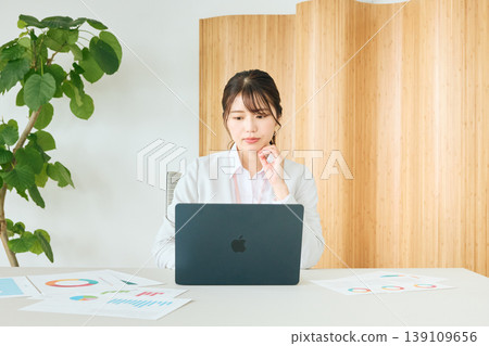 A young female business professional thinking while looking at her laptop in the office; a desk-work style with documents and graphs spread out. A young female business professional thinking while looking at her laptop in the office; a desk-work style with documents and graphs spread out. 139109656