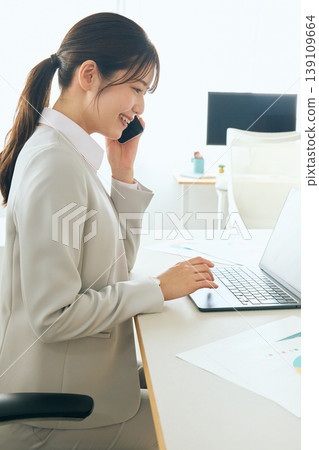 A smiling young Japanese businesswoman is using her laptop while talking on her smartphone in the office. A smiling young Japanese businesswoman is using her laptop while talking on her smartphone in the office. 139109664