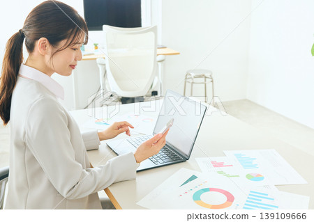 A young female business professional is reviewing graph data using her laptop and smartphone in the office. 139109666