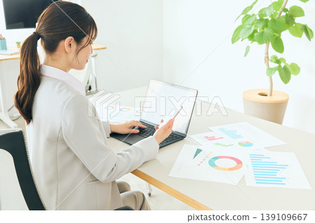 A female business professional and data analyst who uses a laptop and smartphone to review documents in the office. 139109667